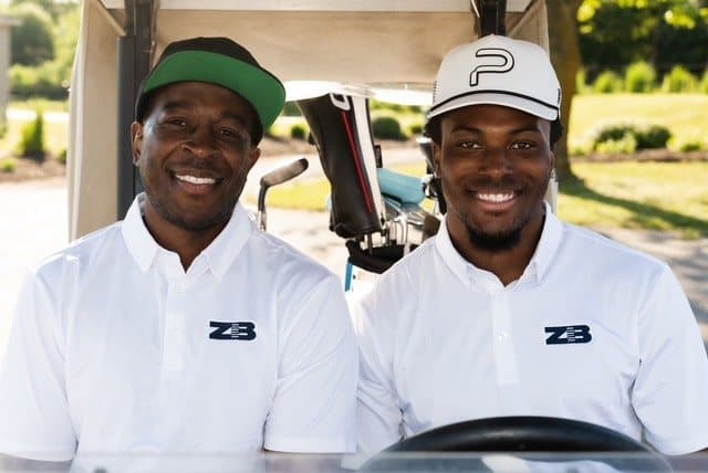 Two golfers in a cart wearing white polos with the ZB logo