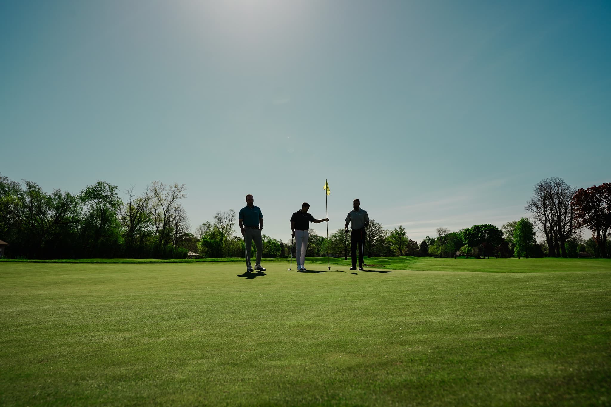 Three golfers by the flag on a parkland green, backlit by the sun
